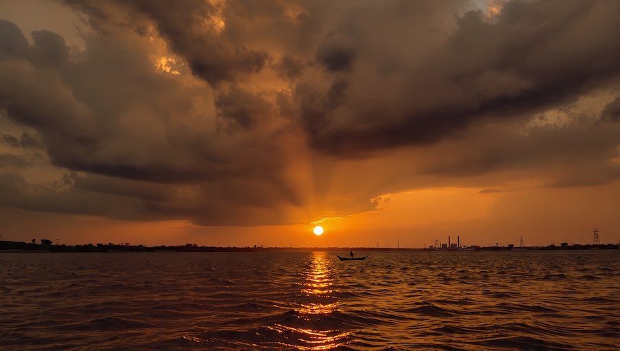 AI-generated sunset image by Meta AI using its own prompt, featuring a silhouette of a boatman on the Damodar River under storm clouds.