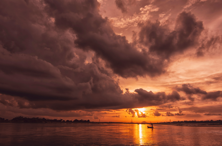 Original real photograph of a dramatic sunset over the Damodar River in Durgapur, showing a silhouetted boat, storm clouds, and industrial structures on the horizon. Image by Arijit Mondal